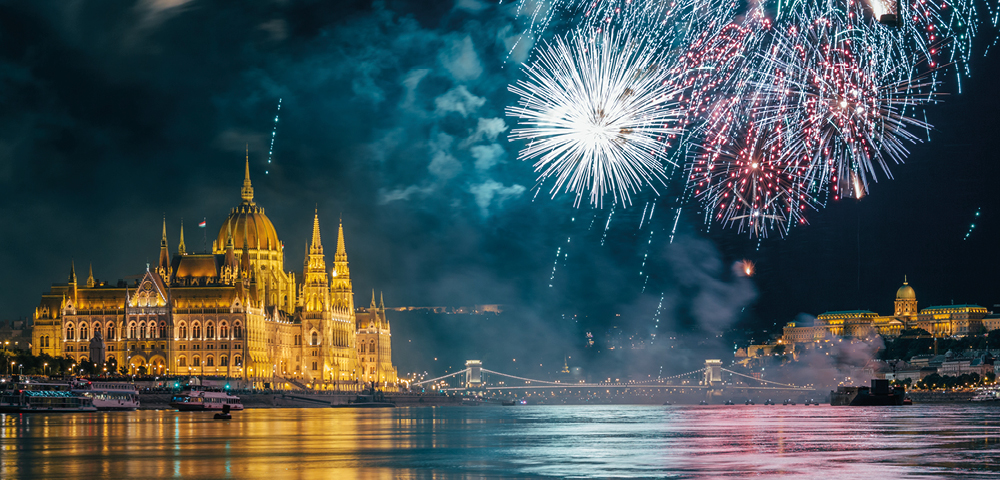 A vibrant fireworks display explodes in the night sky above the illuminated Hungarian Parliament Building and the Danube River.