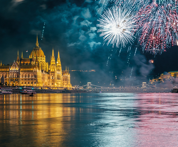 A vibrant fireworks display explodes in the night sky above the illuminated Hungarian Parliament Building and the Danube River.