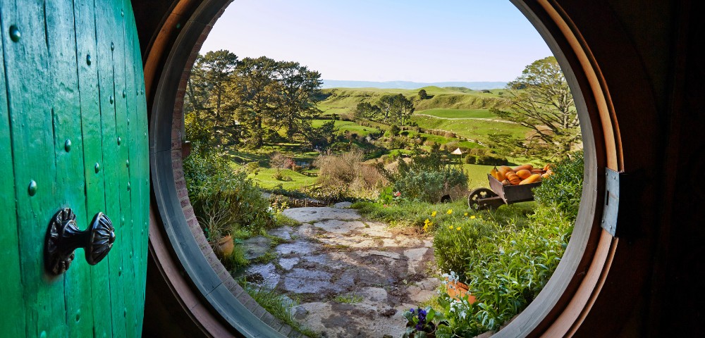 View through a round door reveals a lush, green landscape with rolling hills, trees, and a clear blue sky, evoking a sense of peace and adventure.