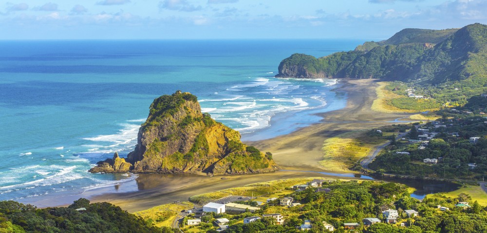 Expansive coastal scene with a prominent rock formation, sandy beaches, and lush greenery. Waves gently crash onshore under a bright blue sky.