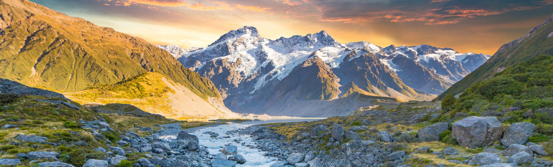 A breathtaking view of snow-capped mountains under a vibrant sunset sky, with a river flowing through a rocky valley, conveying tranquility and awe.