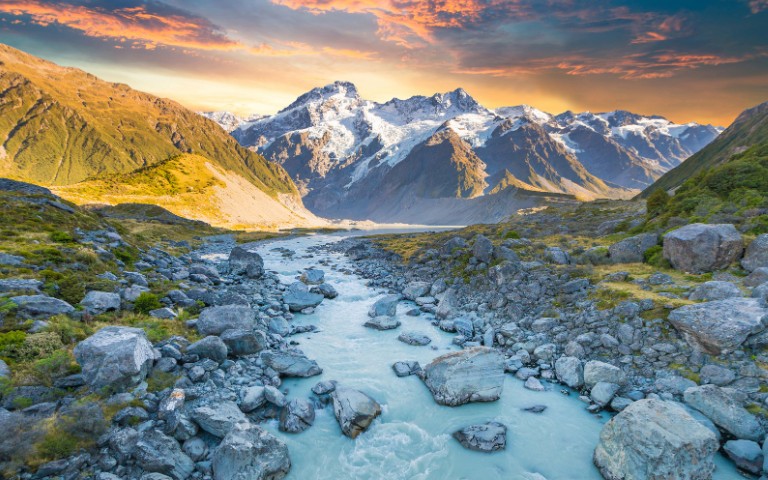 A breathtaking view of snow-capped mountains under a vibrant sunset sky, with a river flowing through a rocky valley, conveying tranquility and awe.