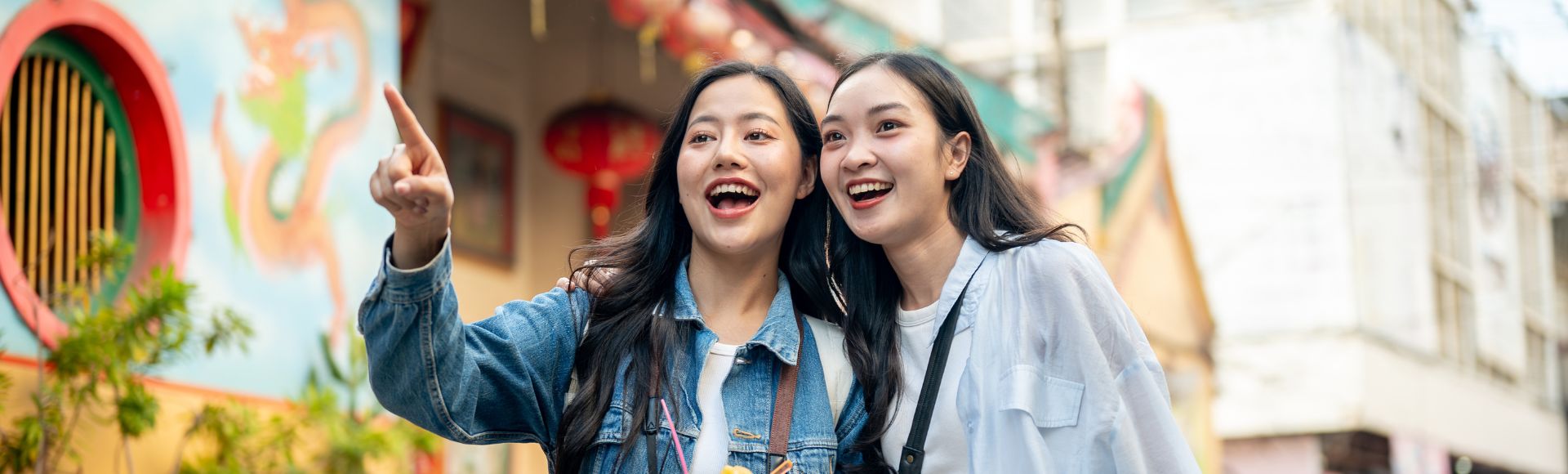 Two smiling women stand outdoors, one pointing excitedly. They are in front of a colorful mural with a red lantern, suggesting a vibrant, lively atmosphere.