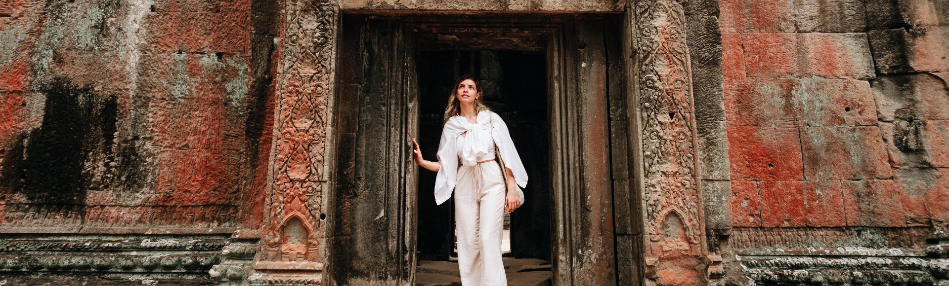 Woman in white outfit stands in an ancient, intricately carved stone doorway, exuding a sense of wonder and exploration amidst weathered architecture.