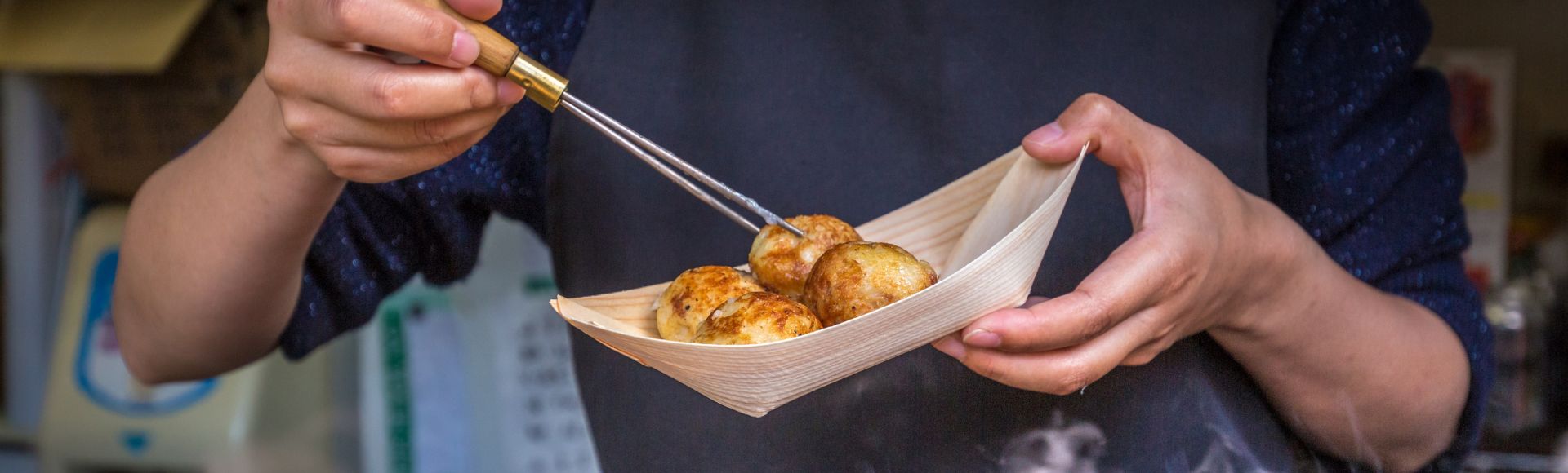 A person holds a wooden tray of takoyaki, using chopsticks to pick one up. The background is blurred, focusing on the food and hands, conveying a street food vibe.