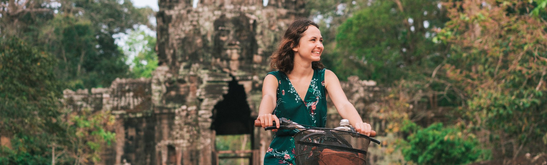 Woman in a green dress smiles while riding a bike in front of an ancient stone archway surrounded by lush greenery, conveying joy and exploration.