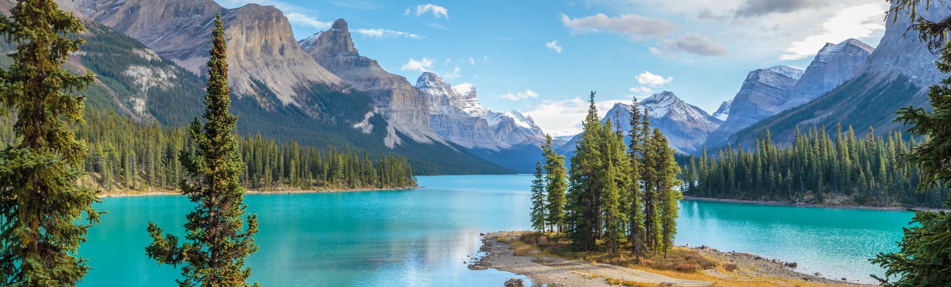 A serene lake with turquoise water, surrounded by dense pine forests and majestic snow-capped mountains under a clear blue sky, evokes tranquility.