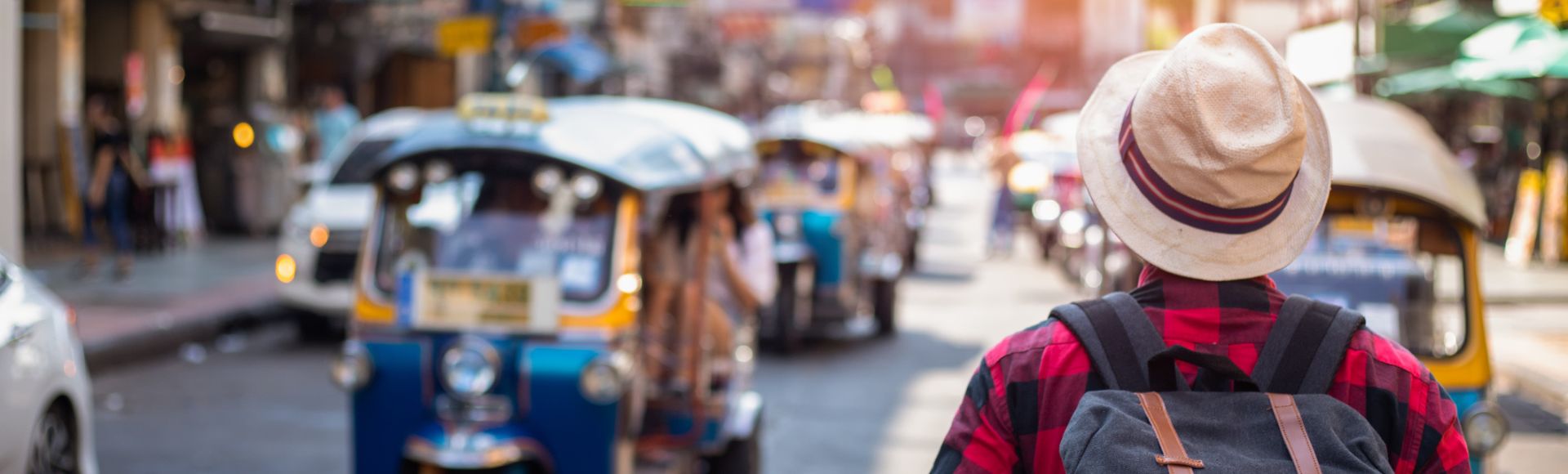 A person in a hat and backpack walks down a bustling street with colorful tuk-tuks under bright sunlight, conveying a sense of travel and adventure.
