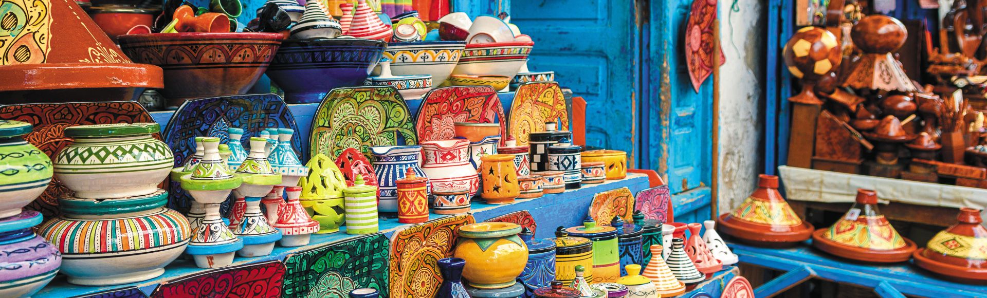 Colorful pottery displayed on a vibrant blue shelf in a market. The ceramic dishes, bowls, and tagines feature intricate patterns, creating a lively, cultural ambiance.