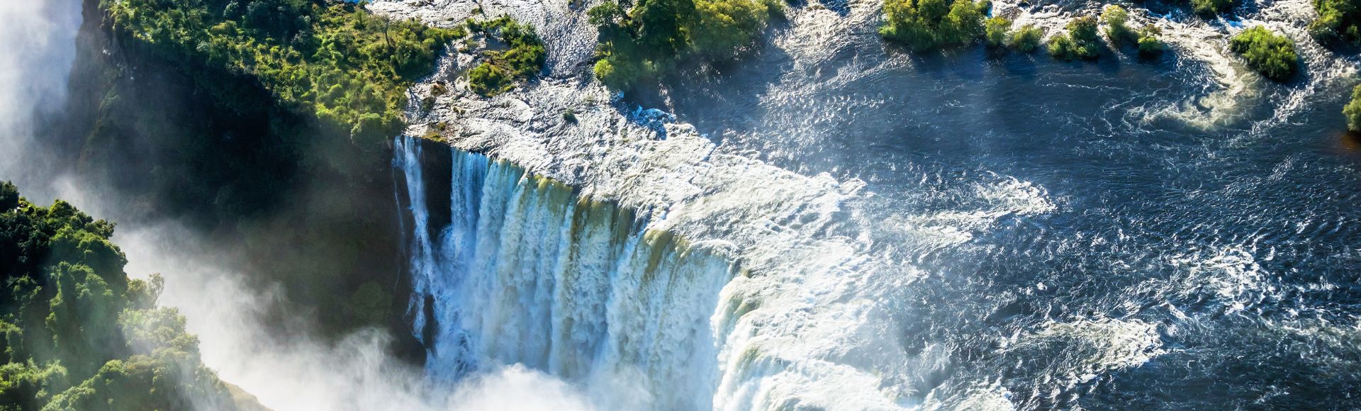 Aerial view of a majestic waterfall cascading over rocky cliffs, surrounded by lush green foliage, with mist rising and water swirling below.