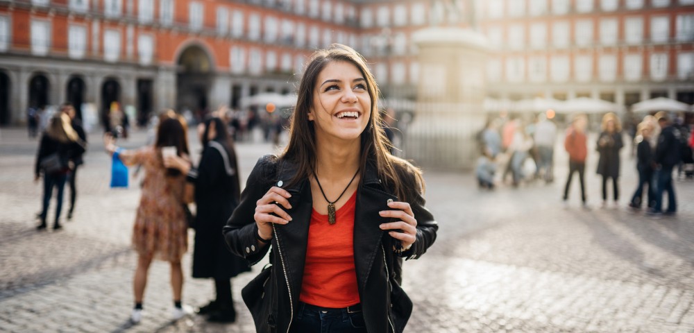 A smiling woman in a black jacket stands in a sunlit plaza, surrounded by blurred people and historic buildings, exuding joy and warmth.