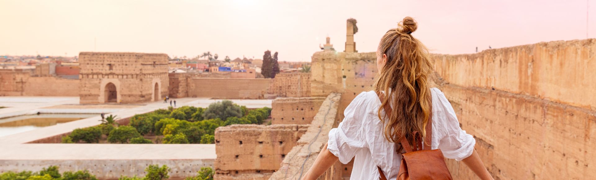 A woman with long hair and a brown backpack stands on an ancient stone wall, overlooking a historic cityscape at sunset, conveying a sense of wonder and exploration.