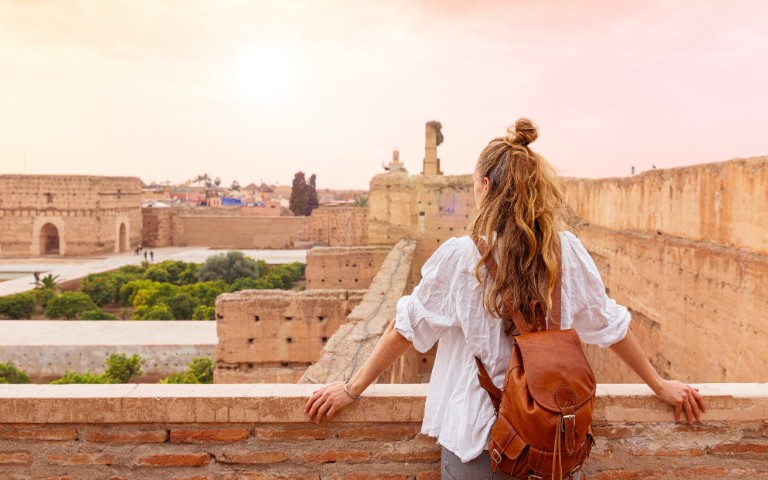 A woman with long hair and a brown backpack stands on an ancient stone wall, overlooking a historic cityscape at sunset, conveying a sense of wonder and exploration.