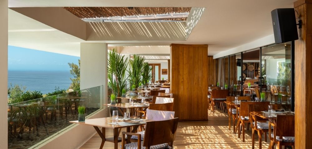 Bright, modern seaside restaurant terrace with wooden furniture, glass railing, green plants, and ocean view under a partial pergola roof.