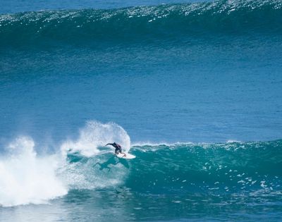 A surfer rides a large blue wave, kicking up spray as they carve across the face of the water.