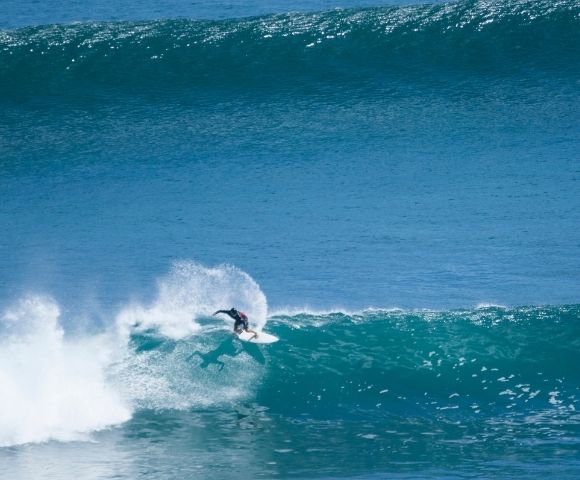 A surfer rides a large blue wave, kicking up spray as they carve across the face of the water.