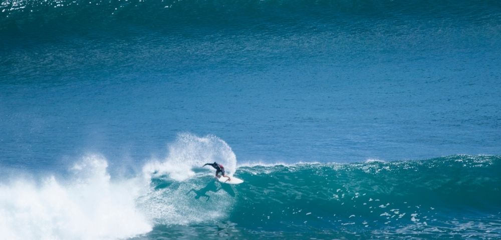 A surfer rides a large blue wave, kicking up spray as they carve across the face of the water.