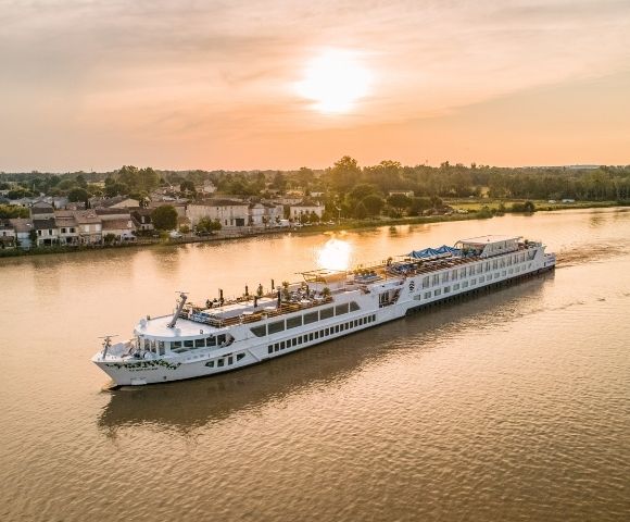 A long, white river cruise ship sails on a wide, calm river at sunset, with a town and trees on the left bank and a line of trees on the right bank.