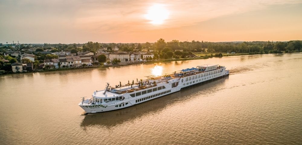 A long, white river cruise ship sails on a wide, calm river at sunset, with a town and trees on the left bank and a line of trees on the right bank.