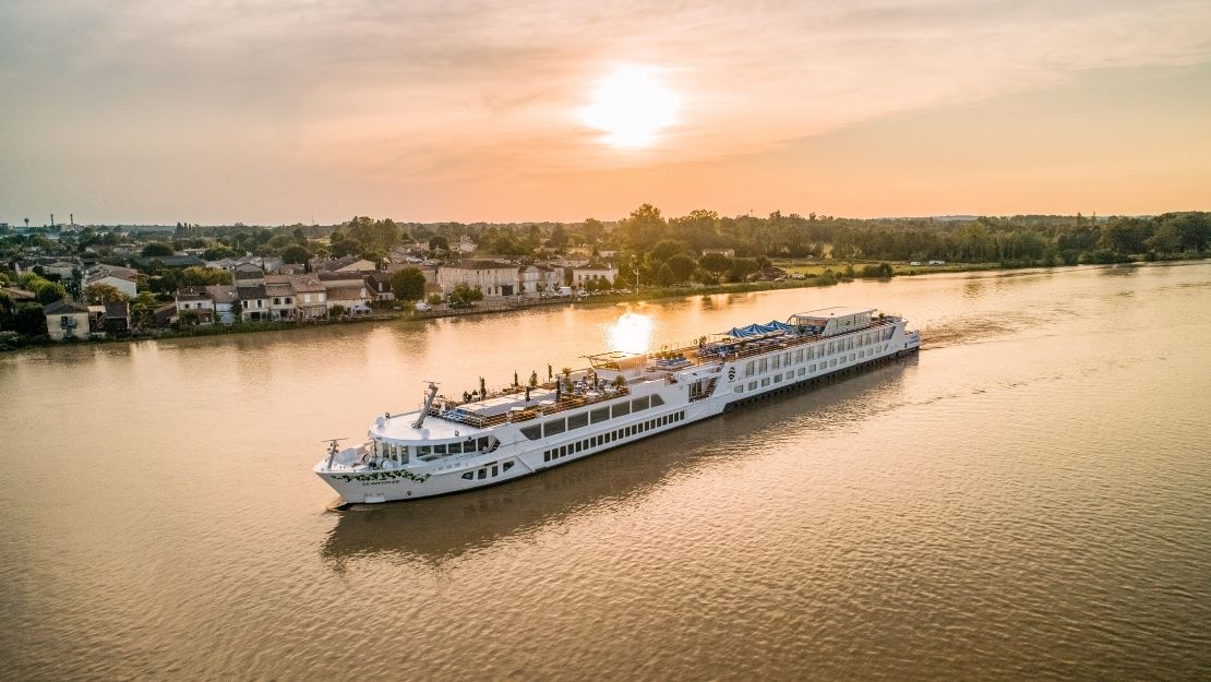A long, white river cruise ship sails on a wide, calm river at sunset, with a town and trees on the left bank and a line of trees on the right bank.