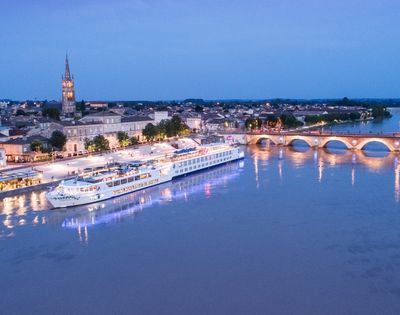 A large white river cruise ship is docked along a waterfront promenade at dusk, with a historic town and arched bridge in the background.