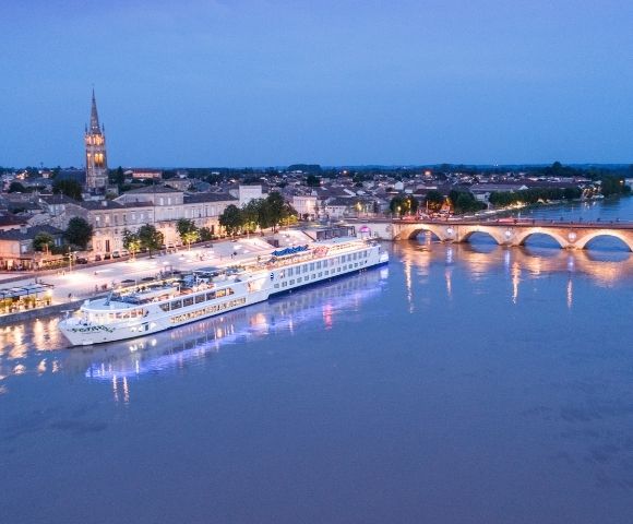 A large white river cruise ship is docked along a waterfront promenade at dusk, with a historic town and arched bridge in the background.