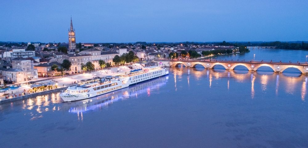 A large white river cruise ship is docked along a waterfront promenade at dusk, with a historic town and arched bridge in the background.