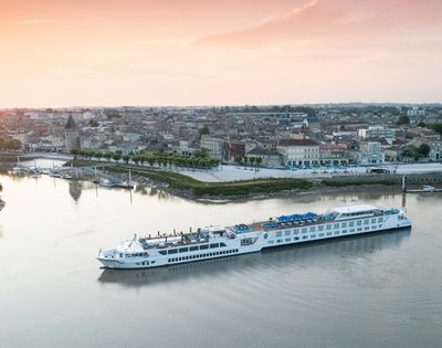 A long white river cruise ship sails on a calm river in front of a picturesque European town with old buildings and a tall church steeple, under a soft sunset sky.