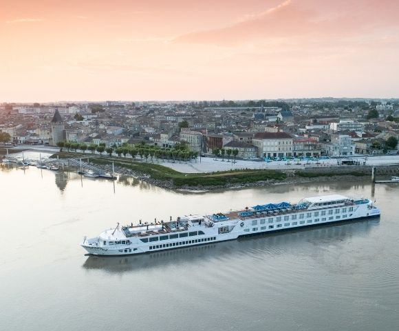 A long white river cruise ship sails on a calm river in front of a picturesque European town with old buildings and a tall church steeple, under a soft sunset sky.