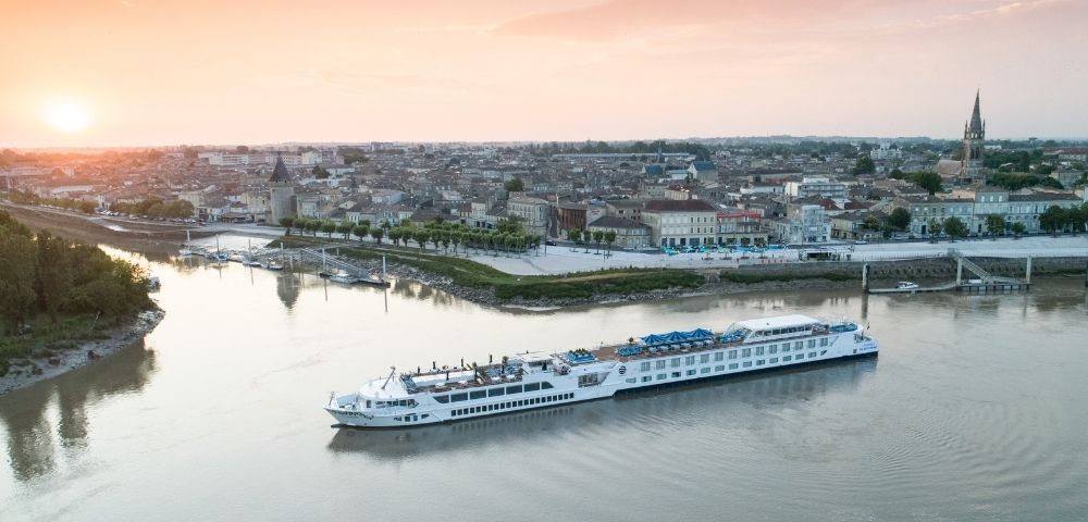 A long white river cruise ship sails on a calm river in front of a picturesque European town with old buildings and a tall church steeple, under a soft sunset sky.