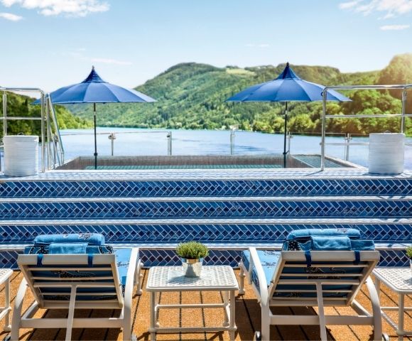 A swimming pool area with lounge chairs and blue umbrellas overlooks a serene lake surrounded by green hills.