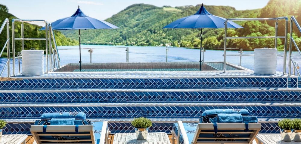 A swimming pool area with lounge chairs and blue umbrellas overlooks a serene lake surrounded by green hills.