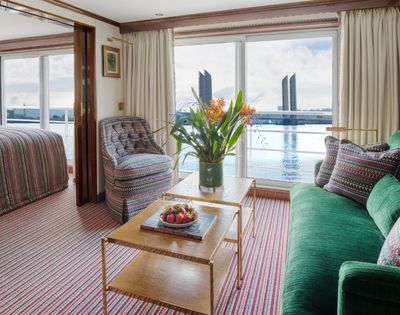 Interior of a river cruise ship cabin with a bed in the background, a patterned armchair, a green sofa with patterned pillows, and a coffee table with strawberries and a book, all overlooking a body of water with modern buildings in the distance.