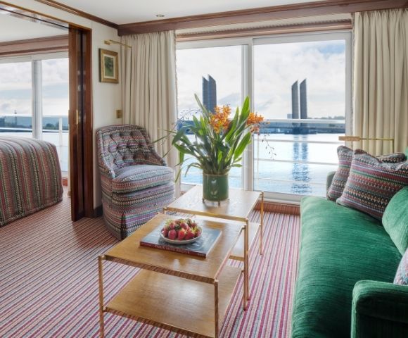 Interior of a river cruise ship cabin with a bed in the background, a patterned armchair, a green sofa with patterned pillows, and a coffee table with strawberries and a book, all overlooking a body of water with modern buildings in the distance.