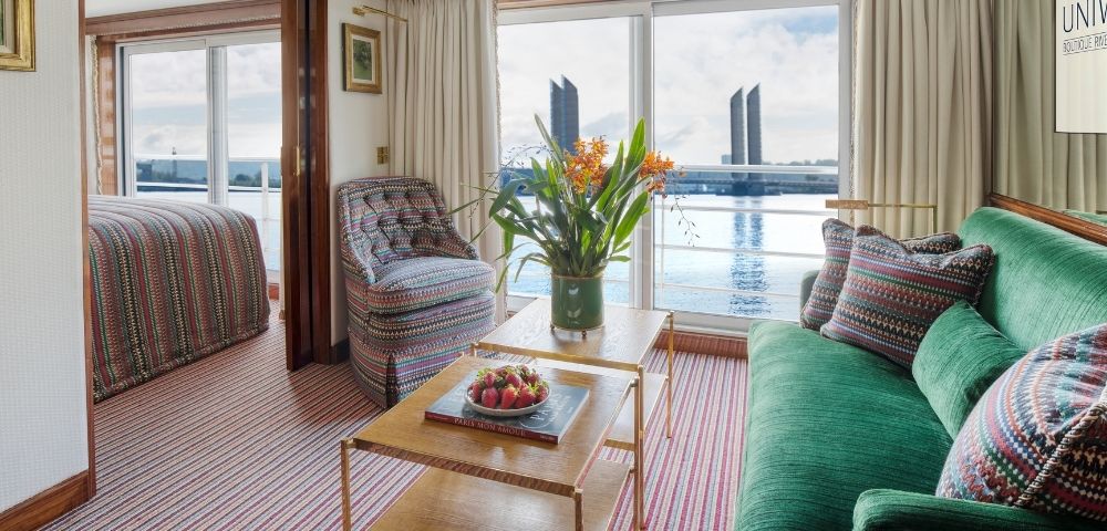 Interior of a river cruise ship cabin with a bed in the background, a patterned armchair, a green sofa with patterned pillows, and a coffee table with strawberries and a book, all overlooking a body of water with modern buildings in the distance.