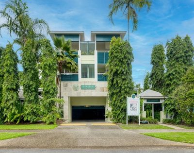 Modern apartment building entrance with manicured trees and palm trees, under a blue sky.