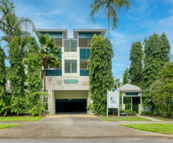 Modern apartment building entrance with manicured trees and palm trees, under a blue sky.