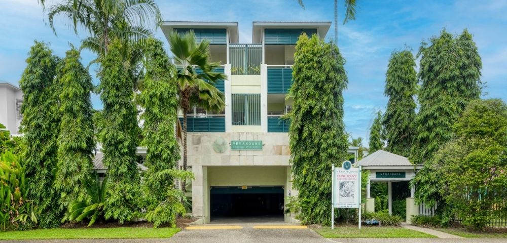 Modern apartment building entrance with manicured trees and palm trees, under a blue sky.