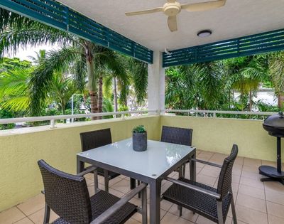 A covered patio with a glass-top dining table and four wicker chairs overlooks lush palm trees, with a grill visible on the right.