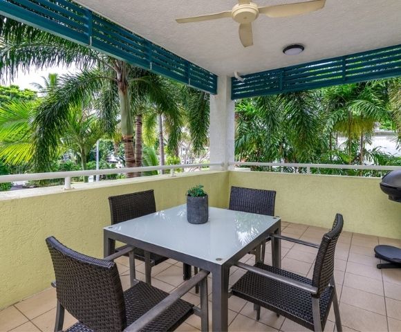 A covered patio with a glass-top dining table and four wicker chairs overlooks lush palm trees, with a grill visible on the right.