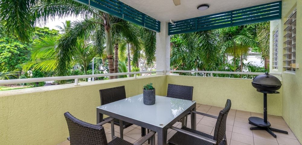 A covered patio with a glass-top dining table and four wicker chairs overlooks lush palm trees, with a grill visible on the right.