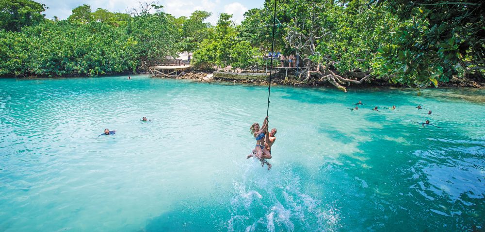 A couple swings from a rope over a crystal-clear turquoise lagoon, surrounded by lush green jungle and other people swimming in the water.