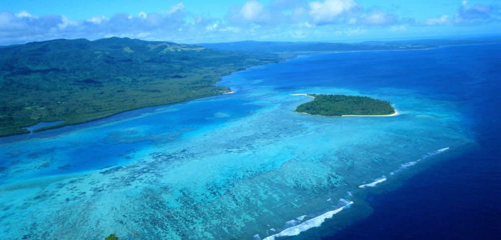 Aerial view of a lush, green island with a sandy beach, surrounded by vibrant turquoise coral reefs and deep blue ocean water.