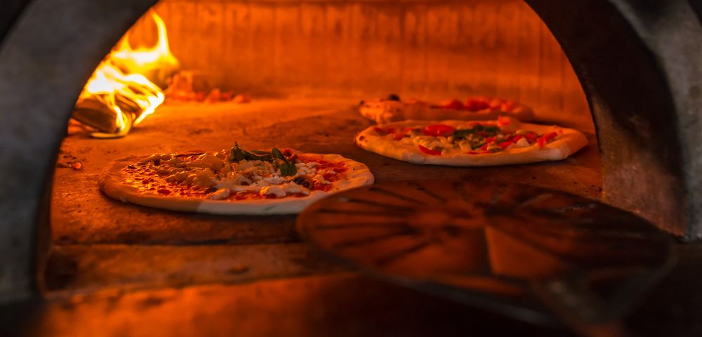 Several pizzas baking in a hot oven with visible flames and a pizza peel in the foreground.