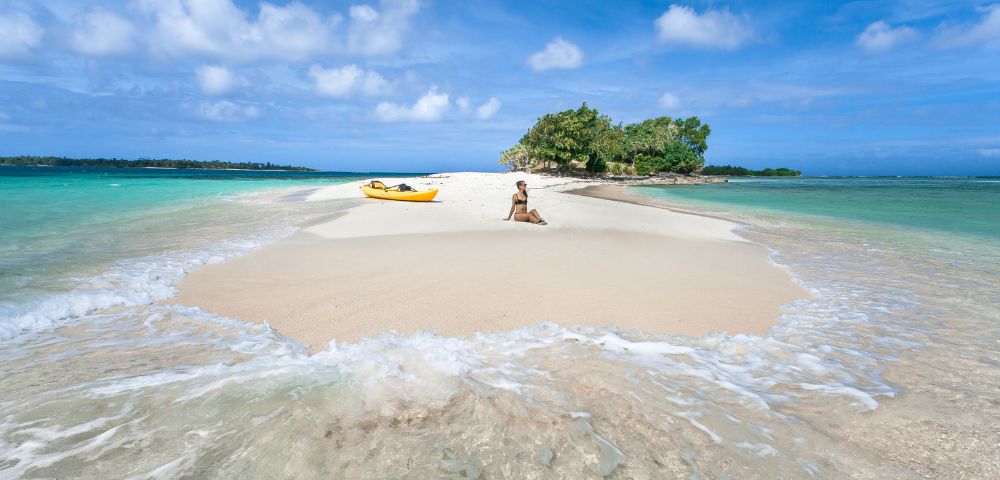 A person in a bikini sits on a white sand beach next to a yellow kayak, with turquoise water and a small island covered in trees in the background under a clear blue sky.