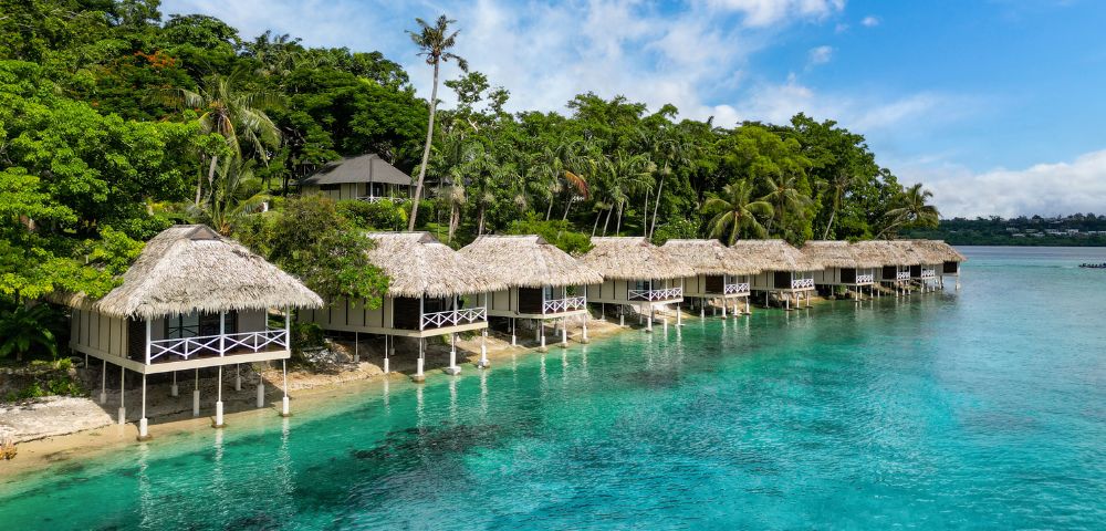 A row of thatched-roof overwater bungalows with white railings and stilts sit on the edge of clear turquoise water, with a lush green hillside and palm trees in the background.