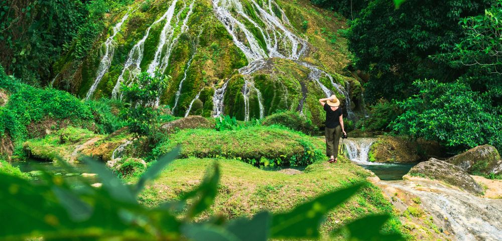 A person wearing a straw hat and dark clothing stands on a grassy ledge, looking towards a lush, moss-covered waterfall cascading down a rocky incline.