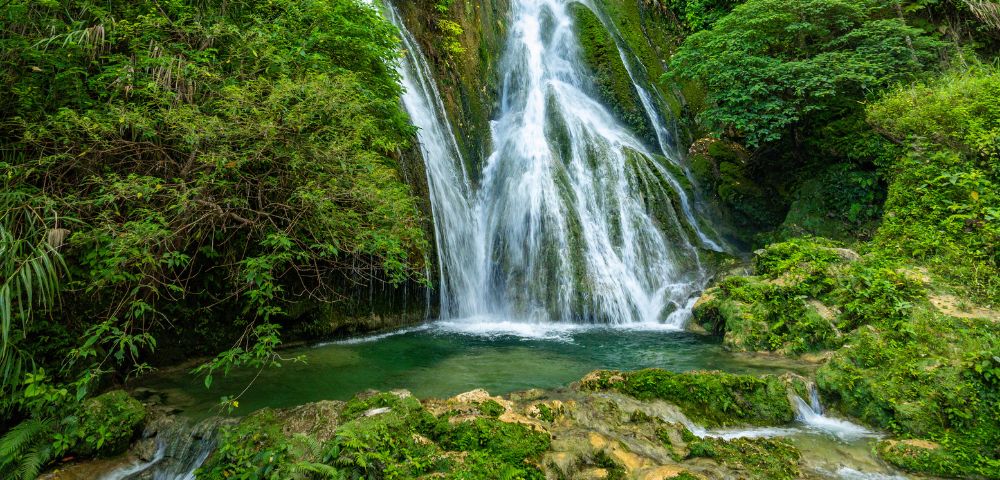 A wide, panoramic view of a multi-tiered waterfall cascading down a mossy rock face surrounded by lush green jungle foliage.