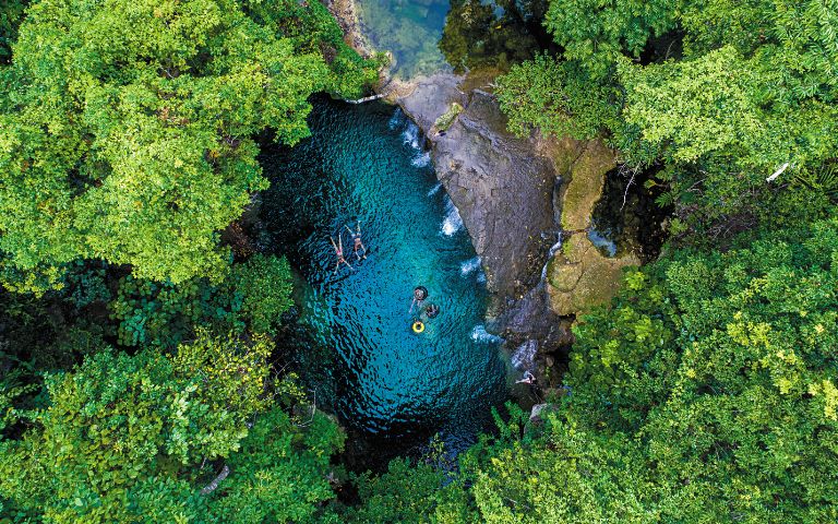 Aerial view of a vibrant turquoise natural pool surrounded by lush green foliage, with several people swimming and floating on tubes.