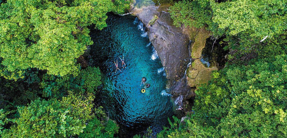 Aerial view of a vibrant turquoise natural pool surrounded by lush green foliage, with several people swimming and floating on tubes.